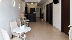 Un vestíbulo de hotel con mostrador de recepción, un área de descanso con sillas y una mesa, y un pasillo con puertas.