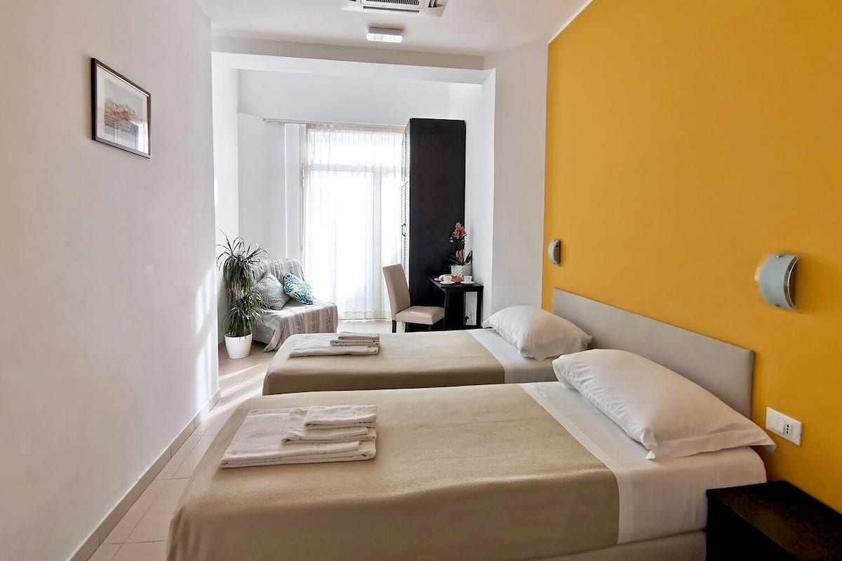 A hotel room with two beds, a desk, a chair, a potted plant, and a window with curtains.