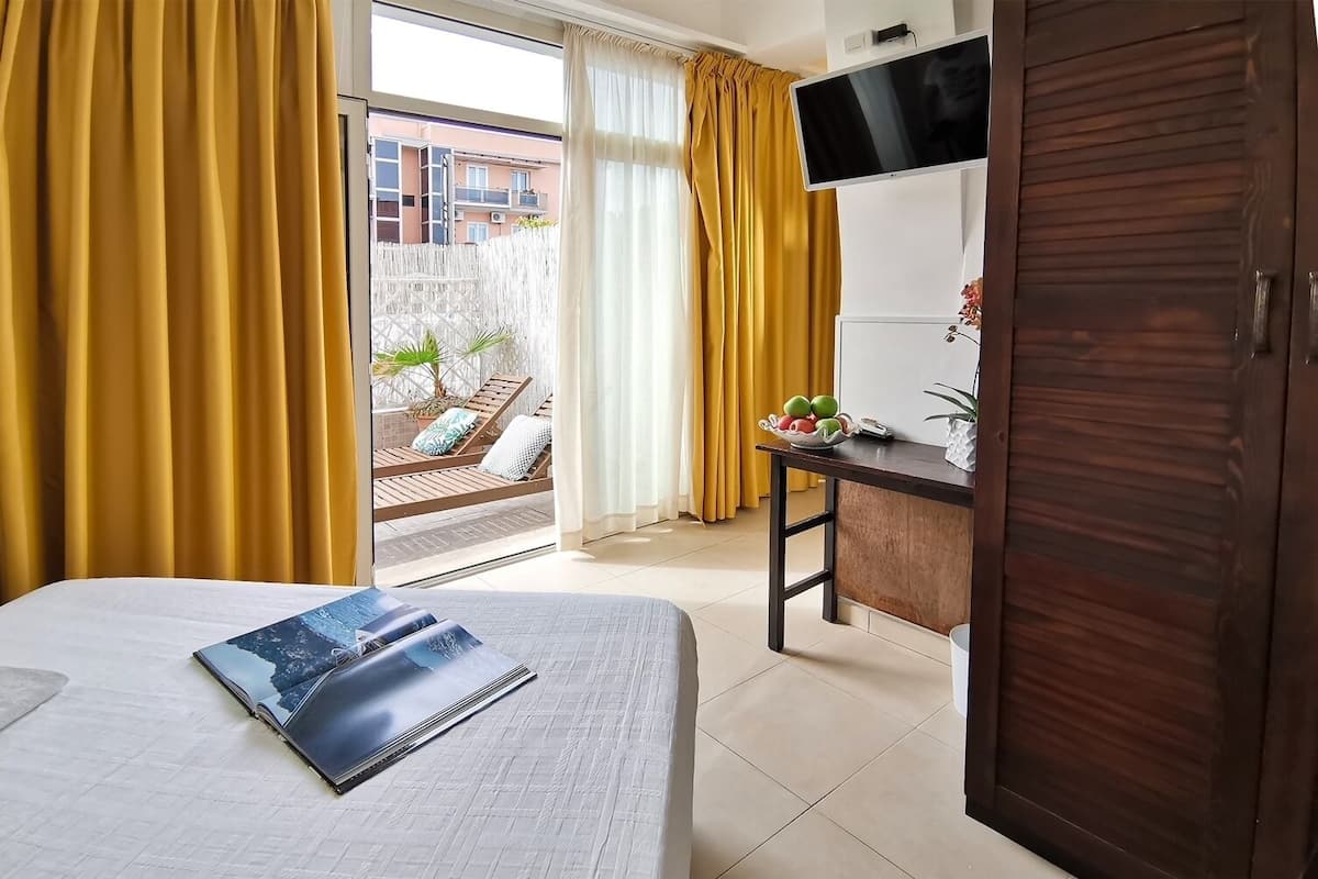 A hotel room with a bed, a desk, a TV, and a balcony with yellow curtains.