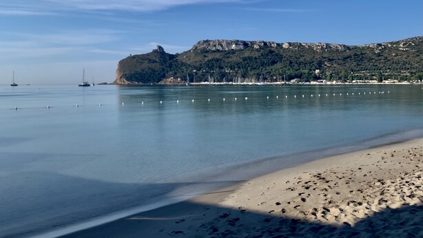 Una playa con aguas tranquilas, una orilla arenosa y una colina rocosa al fondo.