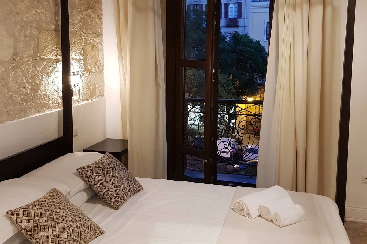 A hotel room with a large bed, two patterned pillows, a nightstand, and a view of a courtyard with a building and trees.