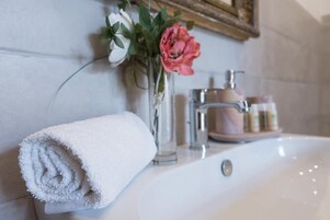 Un bagno con un lavandino bianco, un asciugamano bianco arrotolato e un vaso con fiori.
