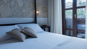 A bedroom with a stone wall, a bed with white linens, patterned pillows, a nightstand with a lamp, and a view through a window with blinds. - La Residenza sul Largo (Cagliari)