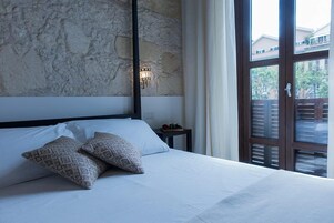 Un dormitorio con pared de piedra, cama con ropa blanca, almohadas estampadas, mesita de noche con lámpara y vistas a través de una ventana con persianas.