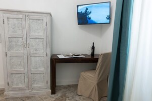 Una habitación con un armario blanco, un escritorio de madera con un libro y una botella, un televisor en la pared y una silla color beige.