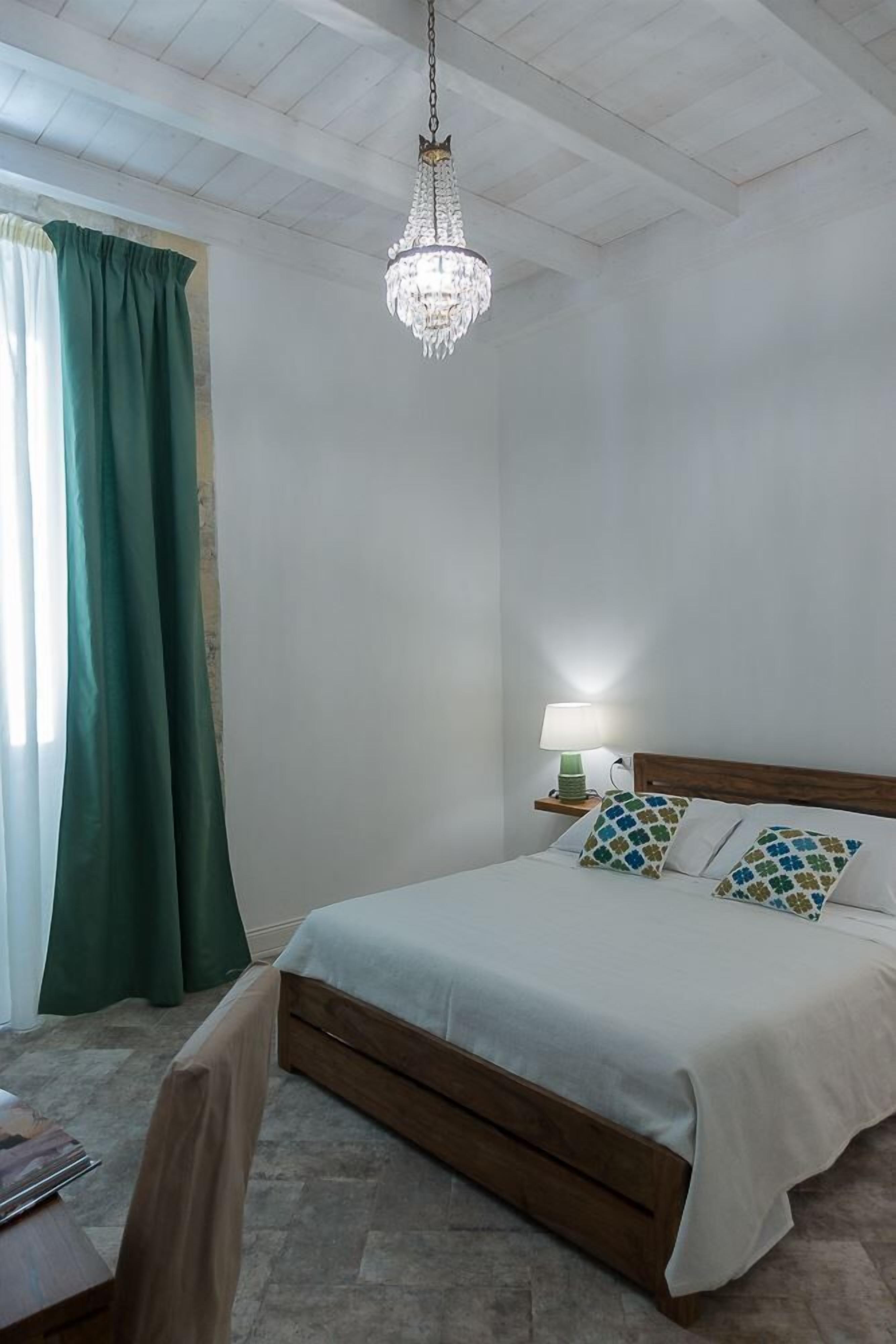 A bedroom with a wooden bed, a chandelier, a bedside table with a lamp, and green curtains.