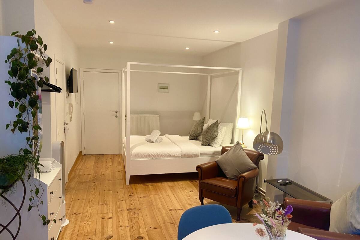A bedroom with a four-poster bed, a wooden floor, a chair, a table with a vase of flowers, and a bedside lamp.