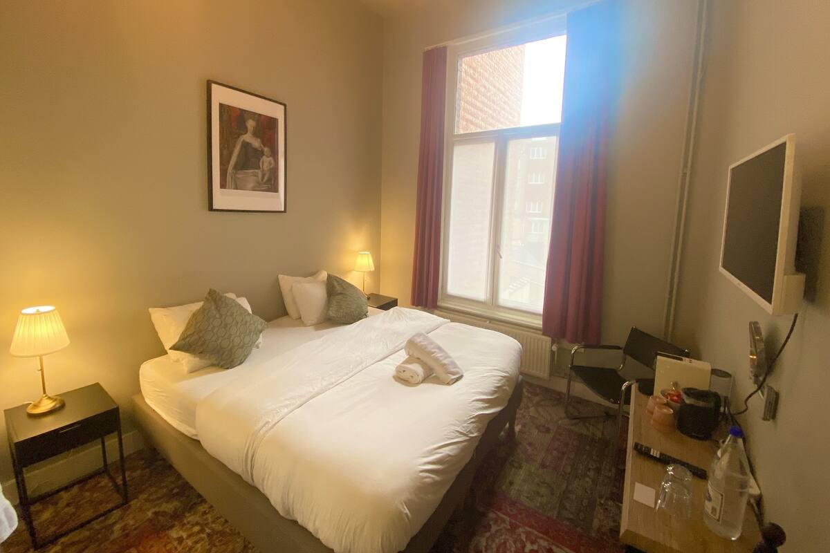 A hotel room with a bed, bedside lamps, a picture on the wall, a window with curtains, a desk with a chair, and a TV mounted on the wall.