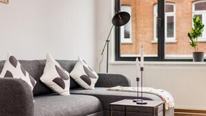 A modern living room with a grey sofa, a small side table, a floor lamp, and a window with a view of a brick building. - Simone's Kitchen B&B (Antwerp)