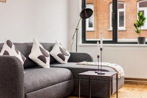 A modern living room with a grey sofa, a small side table, a floor lamp, and a window with a view of a brick building. - Simone's Kitchen B&B (Antwerp)