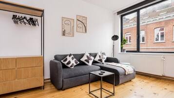 Un salon moderne avec un canapé gris, une petite table et une fenêtre donnant sur un bâtiment en briques.