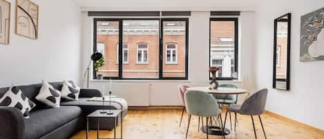 Un salon moderne comprenant un canapé, une table basse et des fauteuils, un coin repas avec une table et des chaises, de grandes fenêtres offrant une vue sur les bâtiments et des œuvres d’art accrochées aux murs.