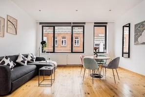 Una sala de estar moderna con un sofá, una mesa de centro y sillas, una zona de comedor con mesa y sillas, ventanas amplias con vistas a edificios, y obras de arte en las paredes.
