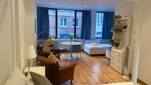 Une chambre d’hôtel moderne avec un plancher en bois, un lit blanc, un canapé en cuir marron, une table à manger ronde et une grande fenêtre avec des rideaux bleus.