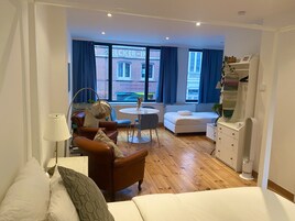 A modern hotel room with a wooden floor, a white bed, a brown leather sofa, a round dining table, and a large window with blue curtains.