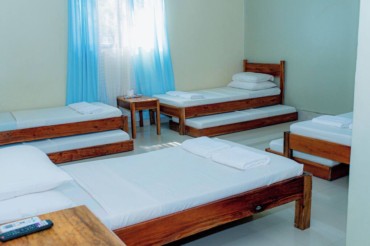 Habitación de hotel con cuatro camas de madera, cada una con ropa blanca y toallas. Hay una pequeña mesa de madera con un control remoto encima.