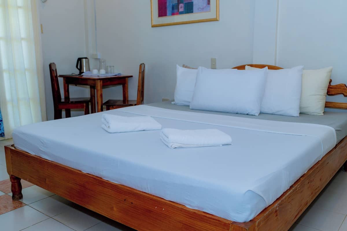 Una cama bien hecha con sábanas y almohadas blancas, un cabecero de madera y una pequeña mesa de madera con una tetera y tazas a un lado.