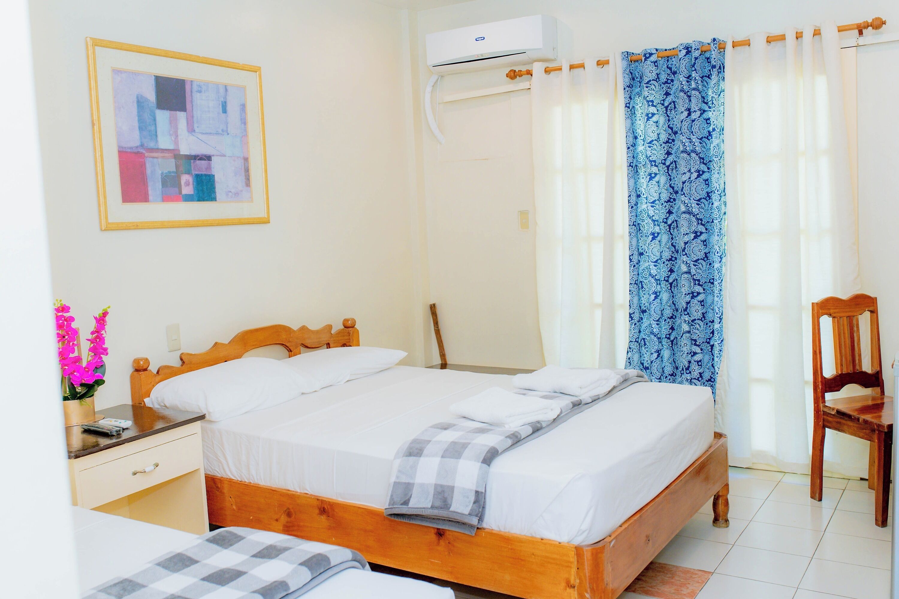 A bedroom with a bed, a nightstand, a chair, a window with curtains, and an air conditioning unit.