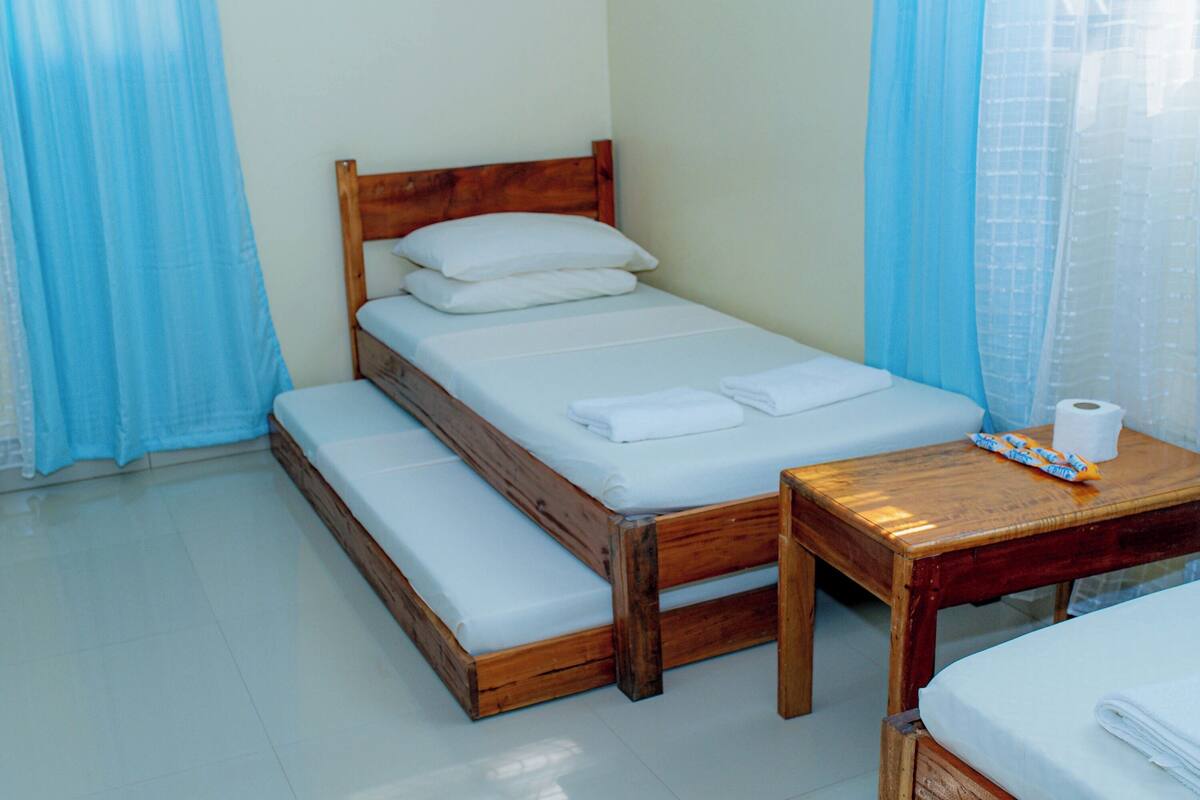 Una cama pequeña y ordenada con cabecera de madera, un banco de madera al pie y una pequeña mesa de madera con un rollo de papel higiénico encima.