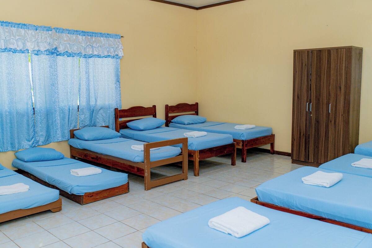 Una habitación con seis camas de madera dispuestas en dos filas, cada una con ropa de cama azul y toallas blancas.
