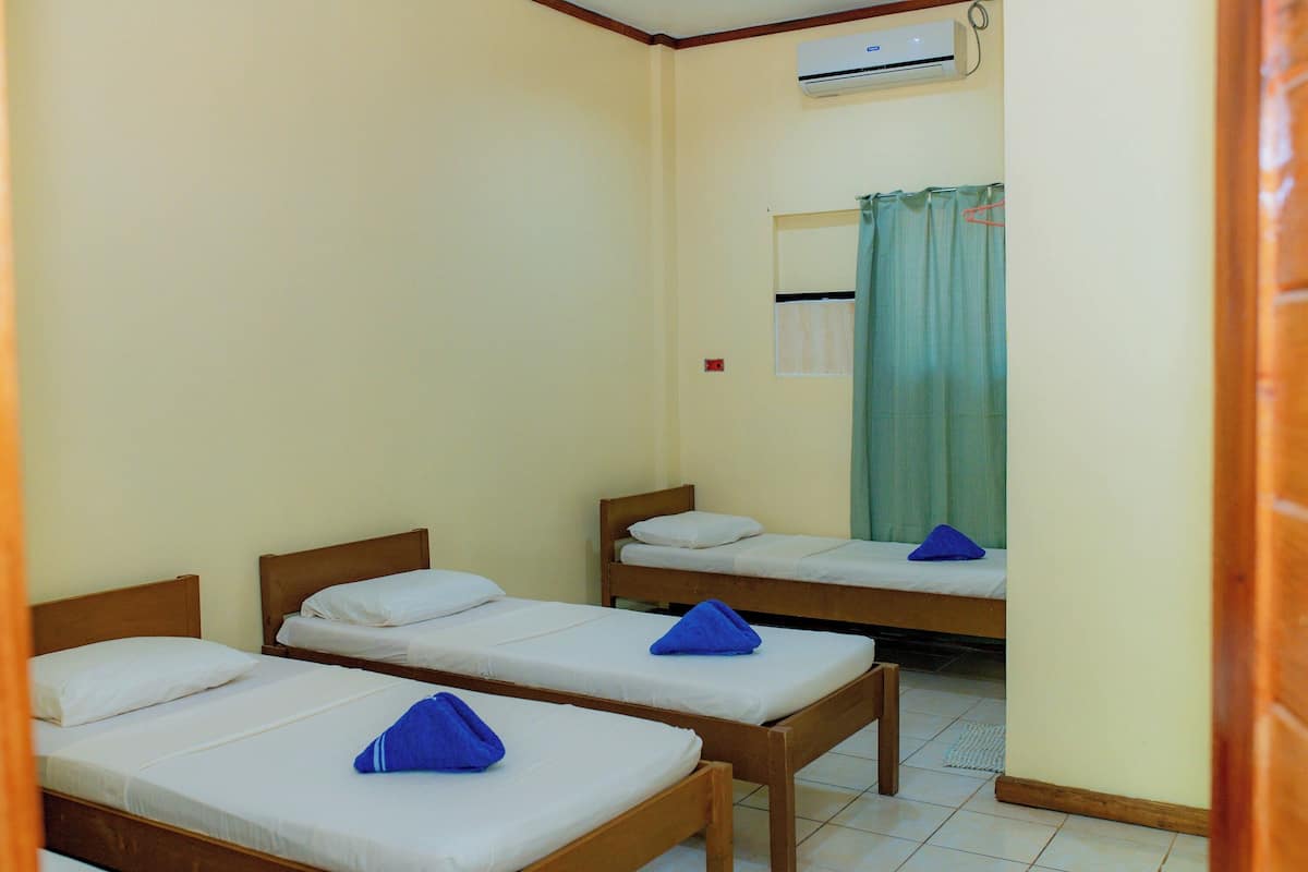 Una habitación con tres camas de madera, cada una con ropa de cama blanca y toallas azules. Un equipo de aire acondicionado está instalado en la pared sobre las camas.