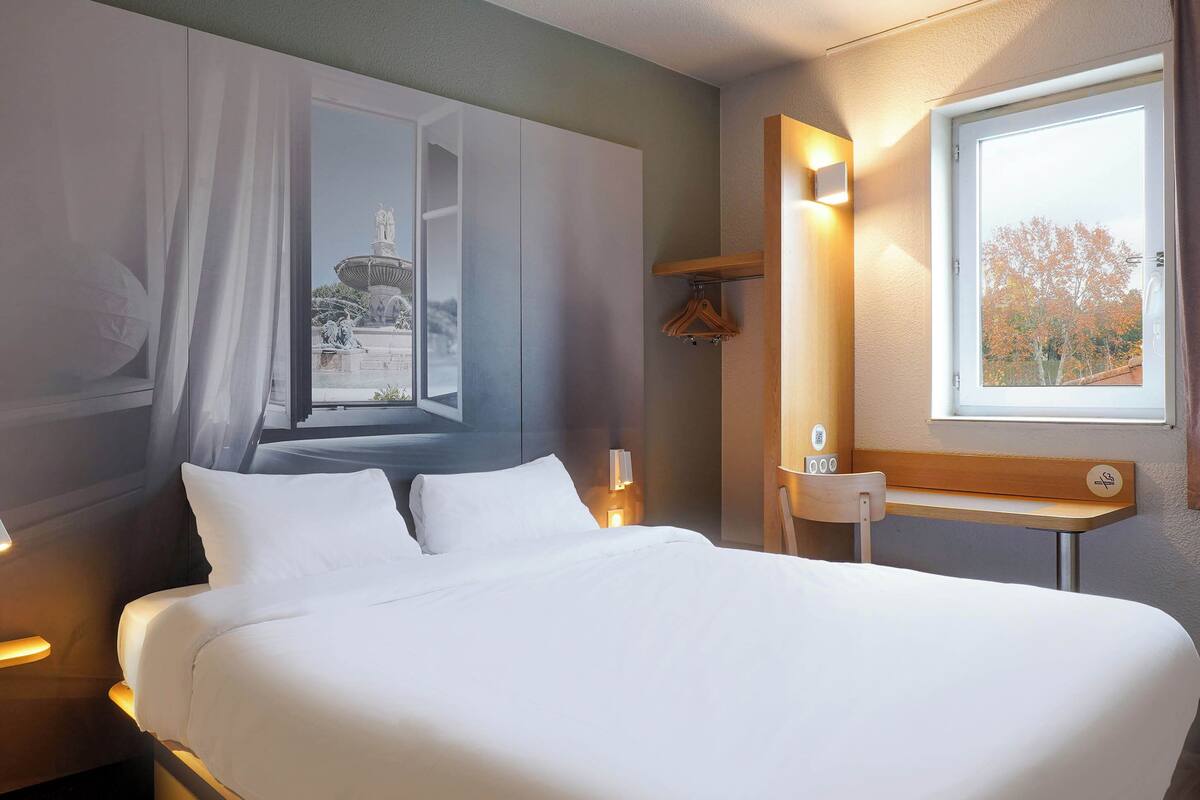 A hotel room with a large bed, a nightstand, a bedside table, a window with a view of a fountain, and a wall-mounted shelf with a mirror and a small table.