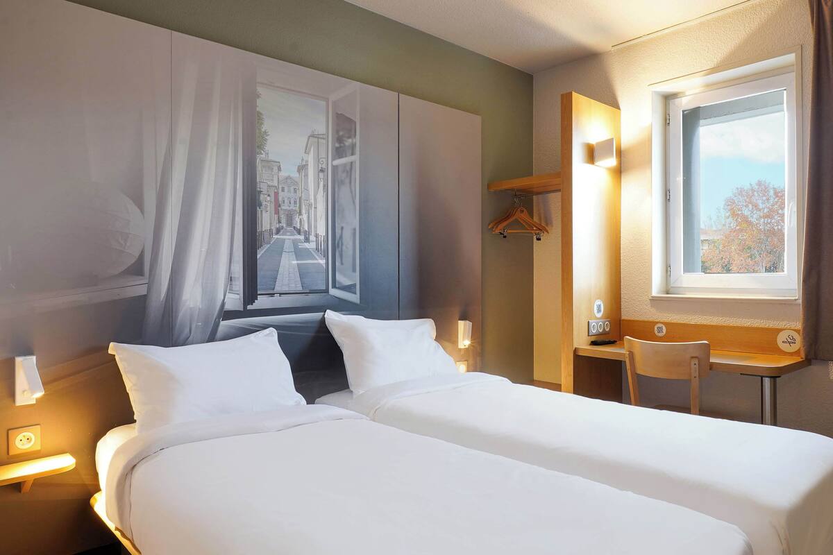 A hotel room with a bed, a desk, a chair, a window with a view, and a wall-mounted light.