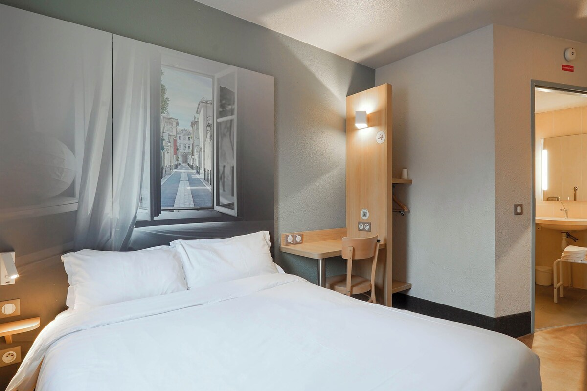 A hotel room with a bed, a desk, a chair, a mirror, and a bathroom visible through an open door.