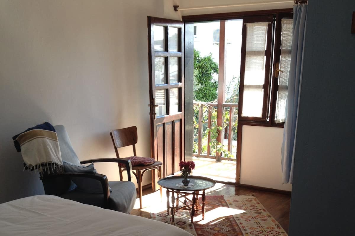 A bedroom with a bed, a chair, a small table with a vase, and a view of the outside through an open door.