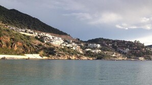 A beach with clear water, pebbly shore, and hillside buildings in the background. - Turk Evi (Kas)