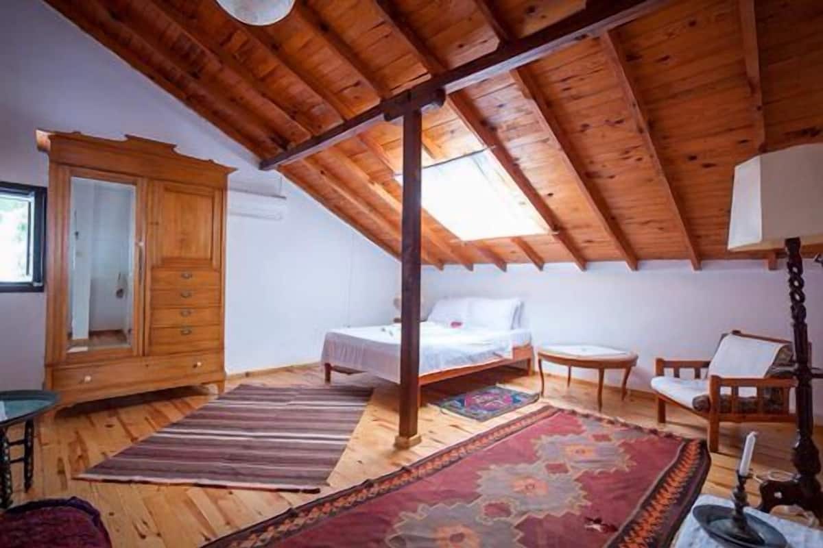 A bedroom with a sloped wooden ceiling, a bed, a small table, a chair, and a large mirror.