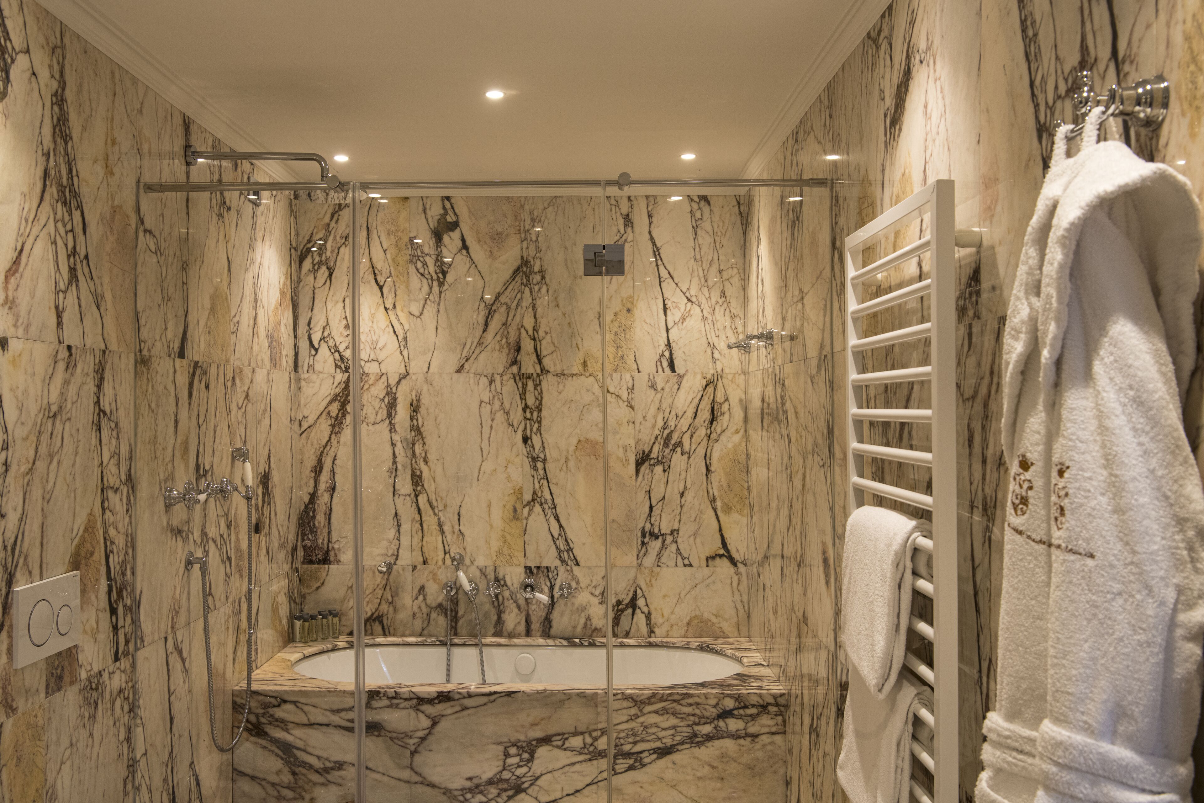 A marble bathroom with a glass-enclosed bathtub, a white towel rack, and a control panel.
