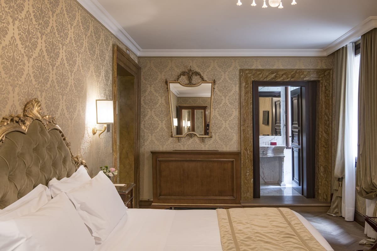 Ett hotellrum med en stor säng, en sänggavel i trä, en spegel och utsikt över badrummet genom en öppen dörr.