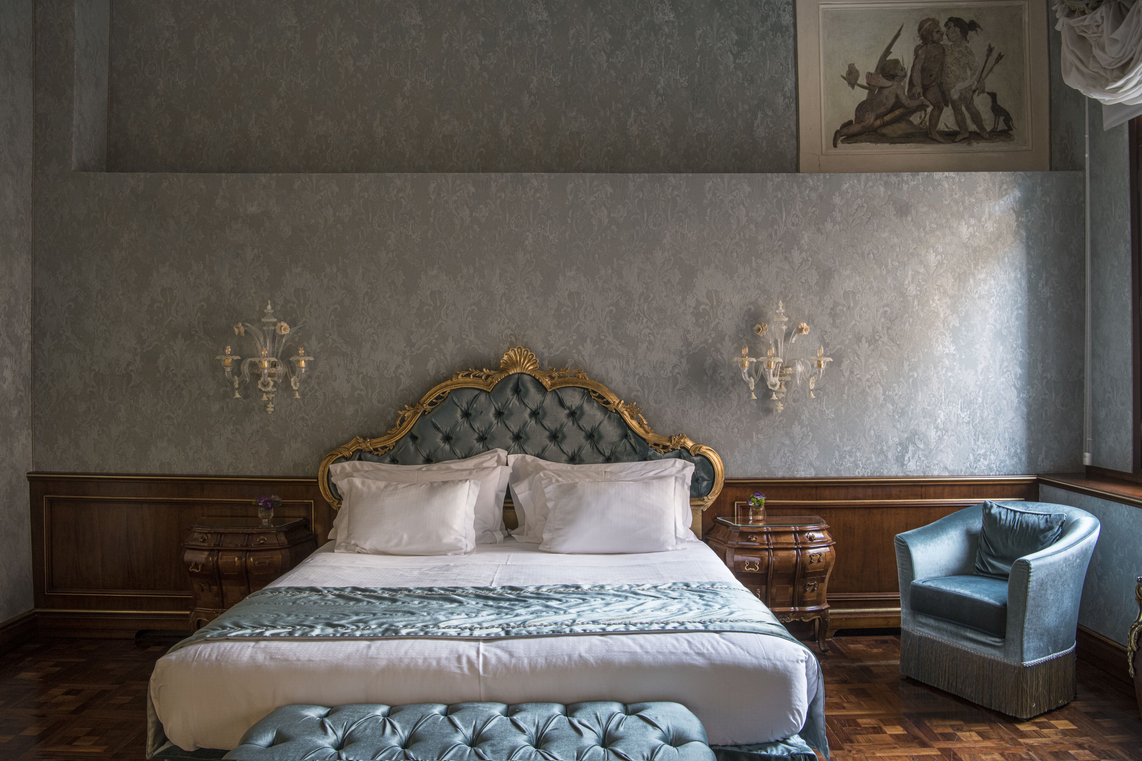 Ein Schlafzimmer mit einem großen Bett, einem gesteppten Kopfteil, zwei Nachttischen, einem Sessel und einem gerahmten Kunstwerk an der Wand.