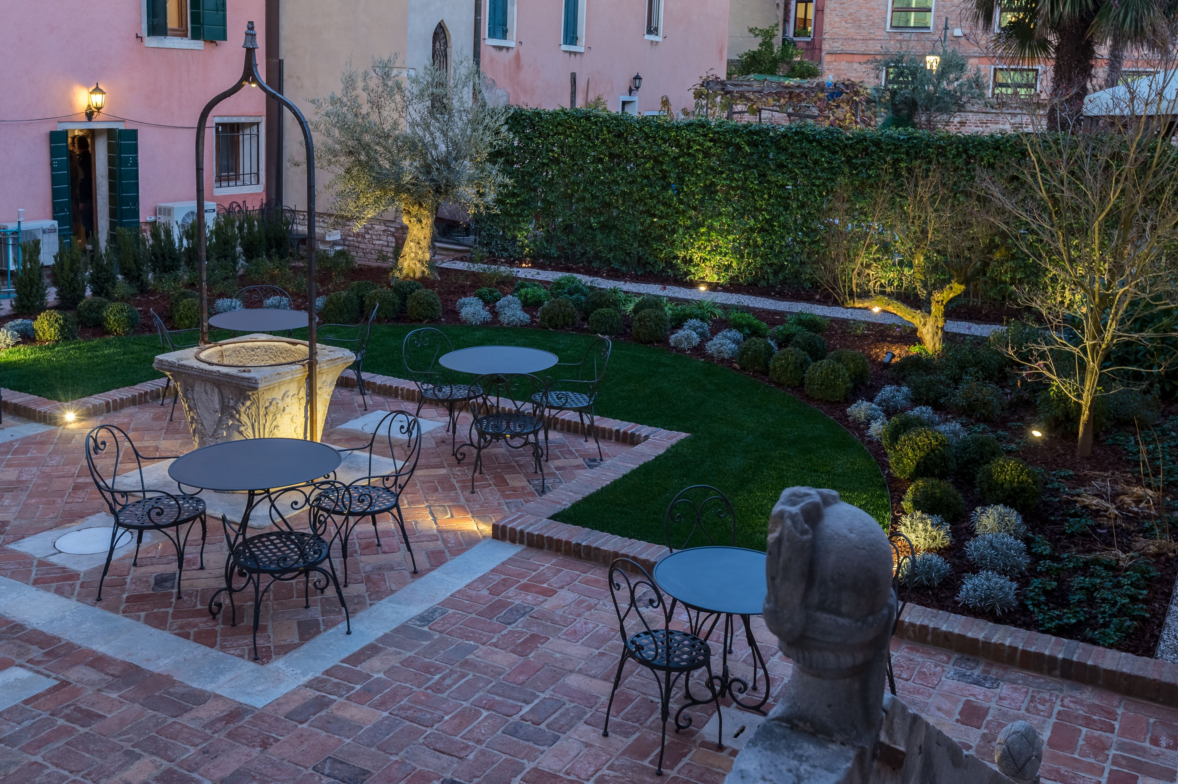 Um pátio com terraço de tijolos, mobiliário de exterior e um jardim bem cuidado, com sebes e árvores.