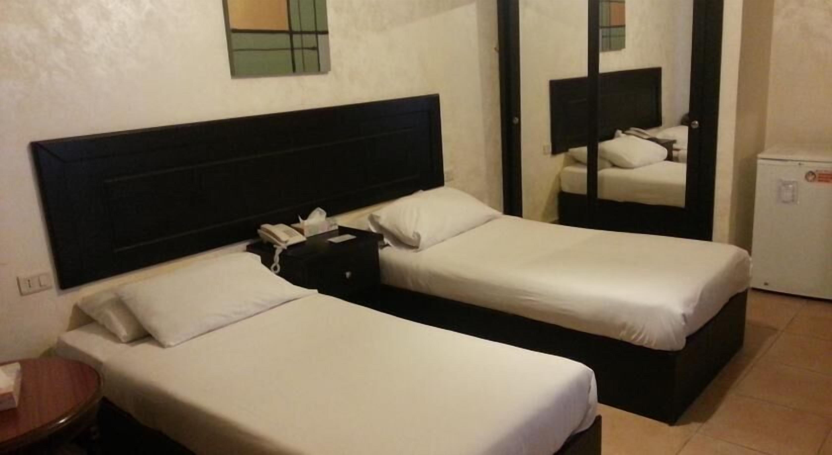 A hotel room with two single beds, a nightstand with a phone, and a small refrigerator.