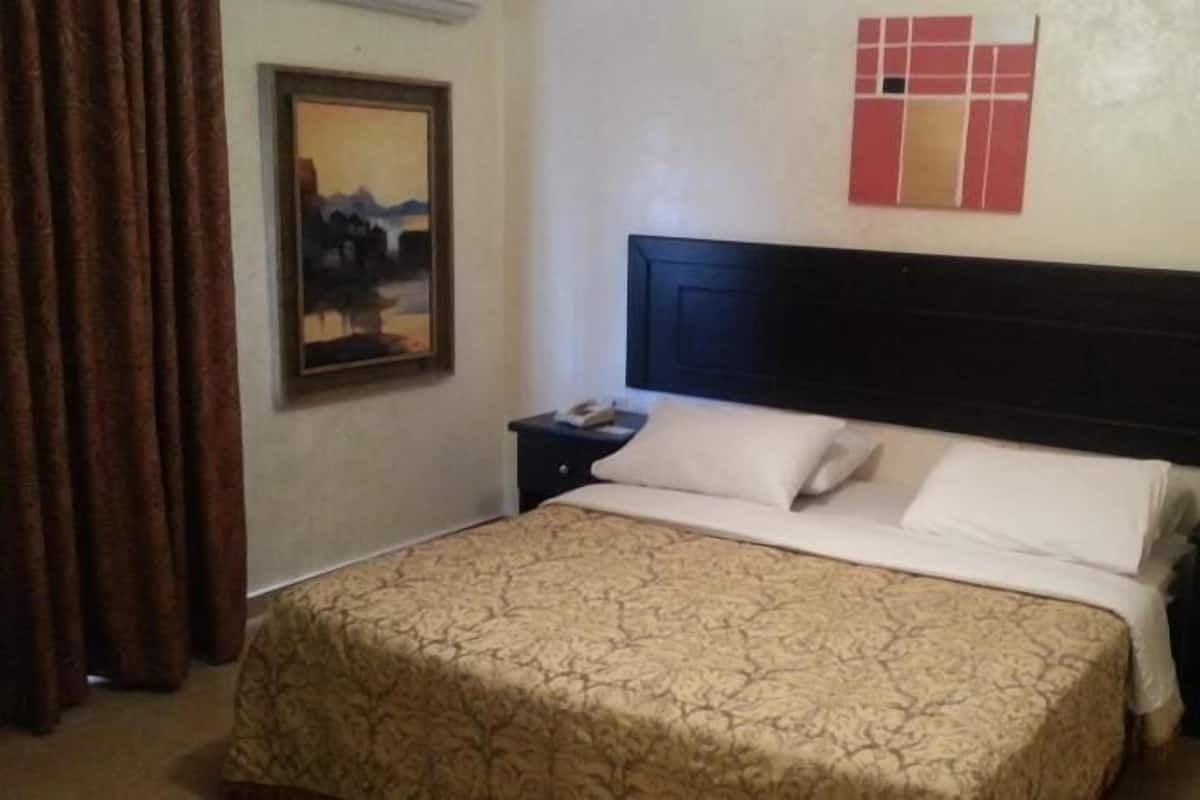 A hotel room with a bed, a nightstand, a painting on the wall, and a window with curtains.