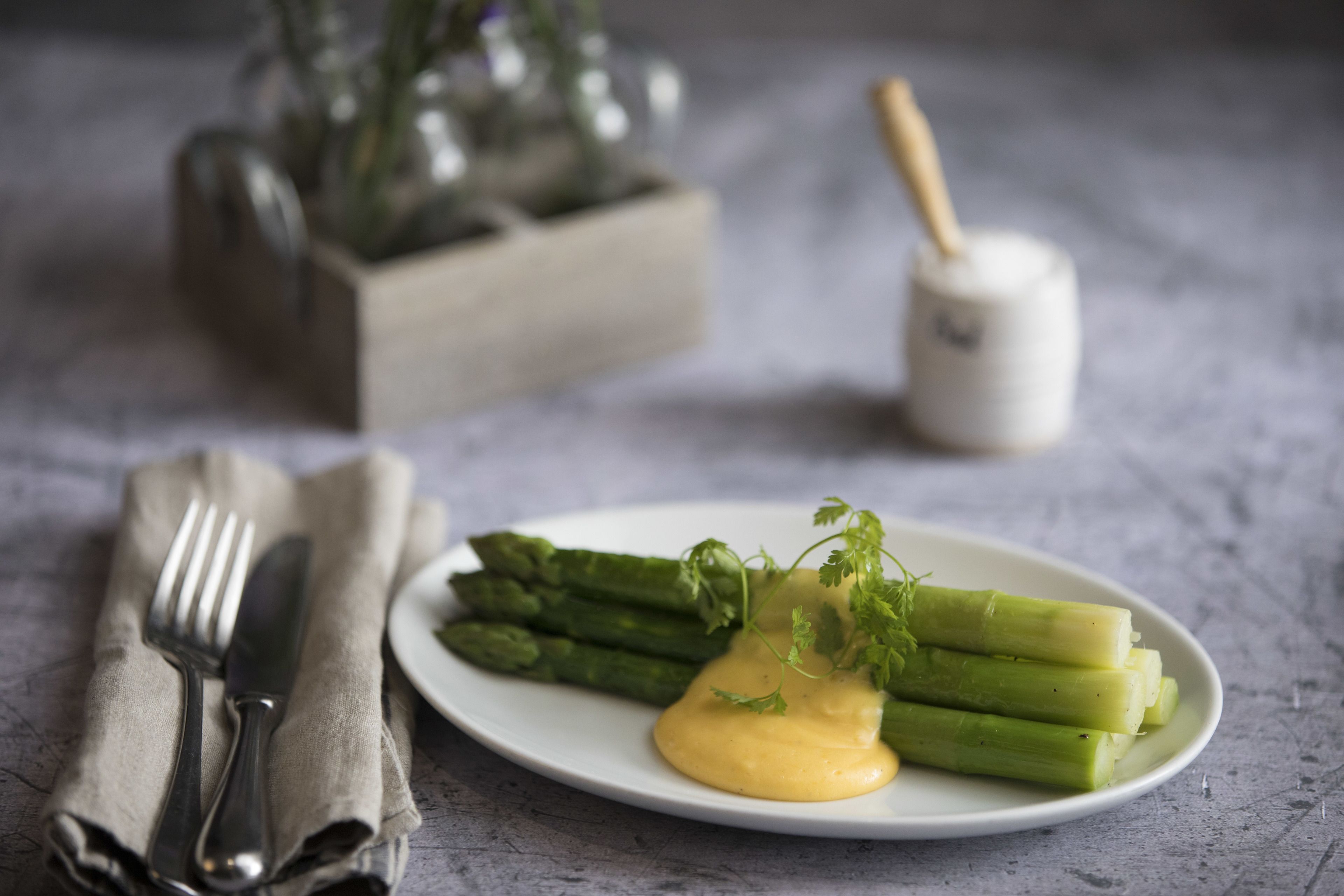 A plate of asparagus with a creamy sauce and a sprig of parsley, a fork and knife on a napkin, and a salt and pepper shaker in the background.