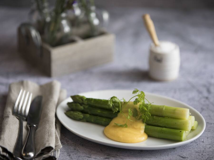 A plate of asparagus with a creamy sauce and a sprig of parsley, a fork and knife on a napkin, and a salt and pepper shaker in the background.