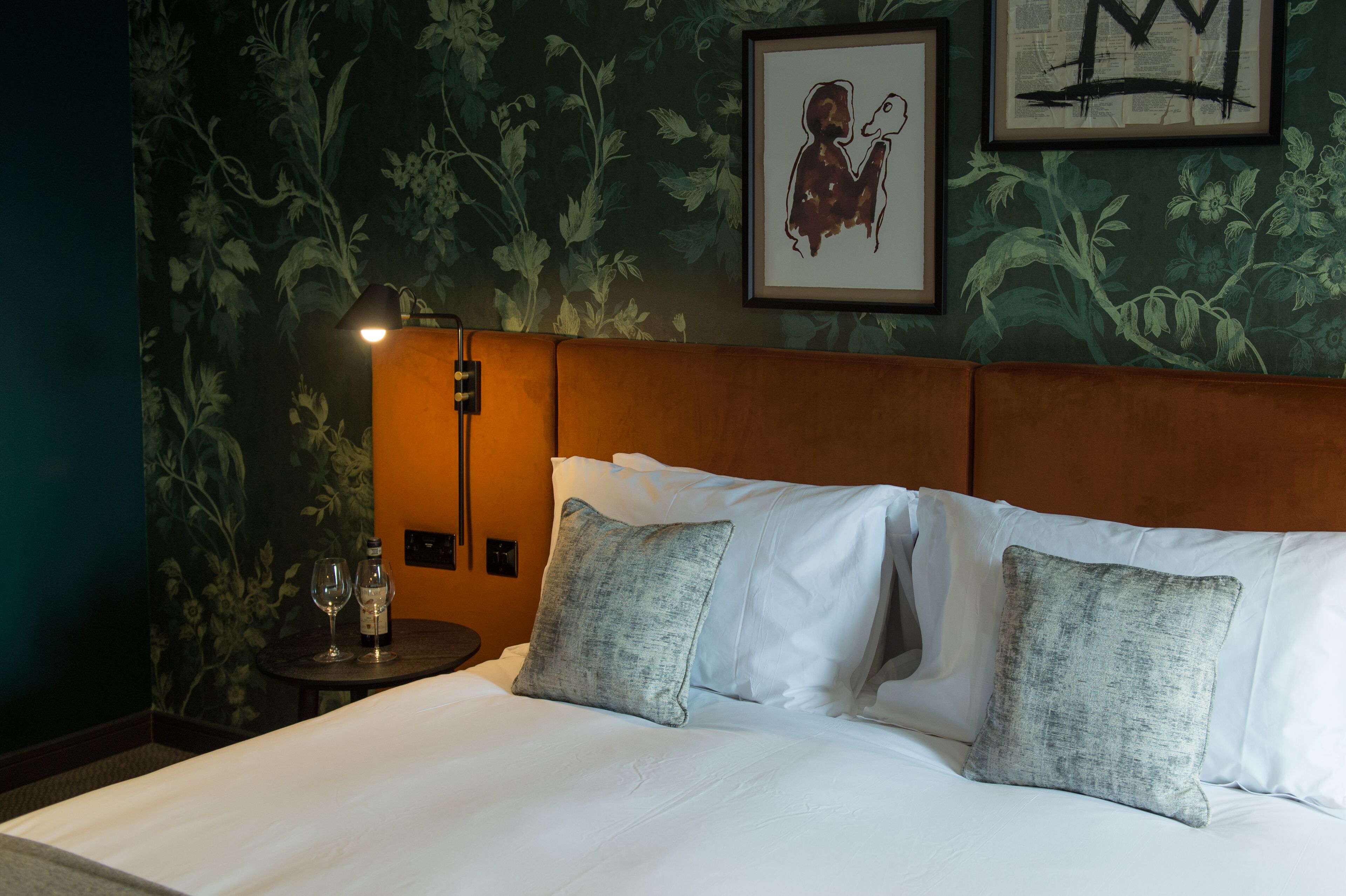A neatly made bed with white linens and two grey pillows, a bedside table with a bottle and glasses, and a framed artwork on the wall.
