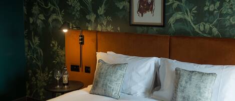 A neatly made bed with white linens and two grey pillows, a bedside table with a bottle and glasses, and a framed artwork on the wall.