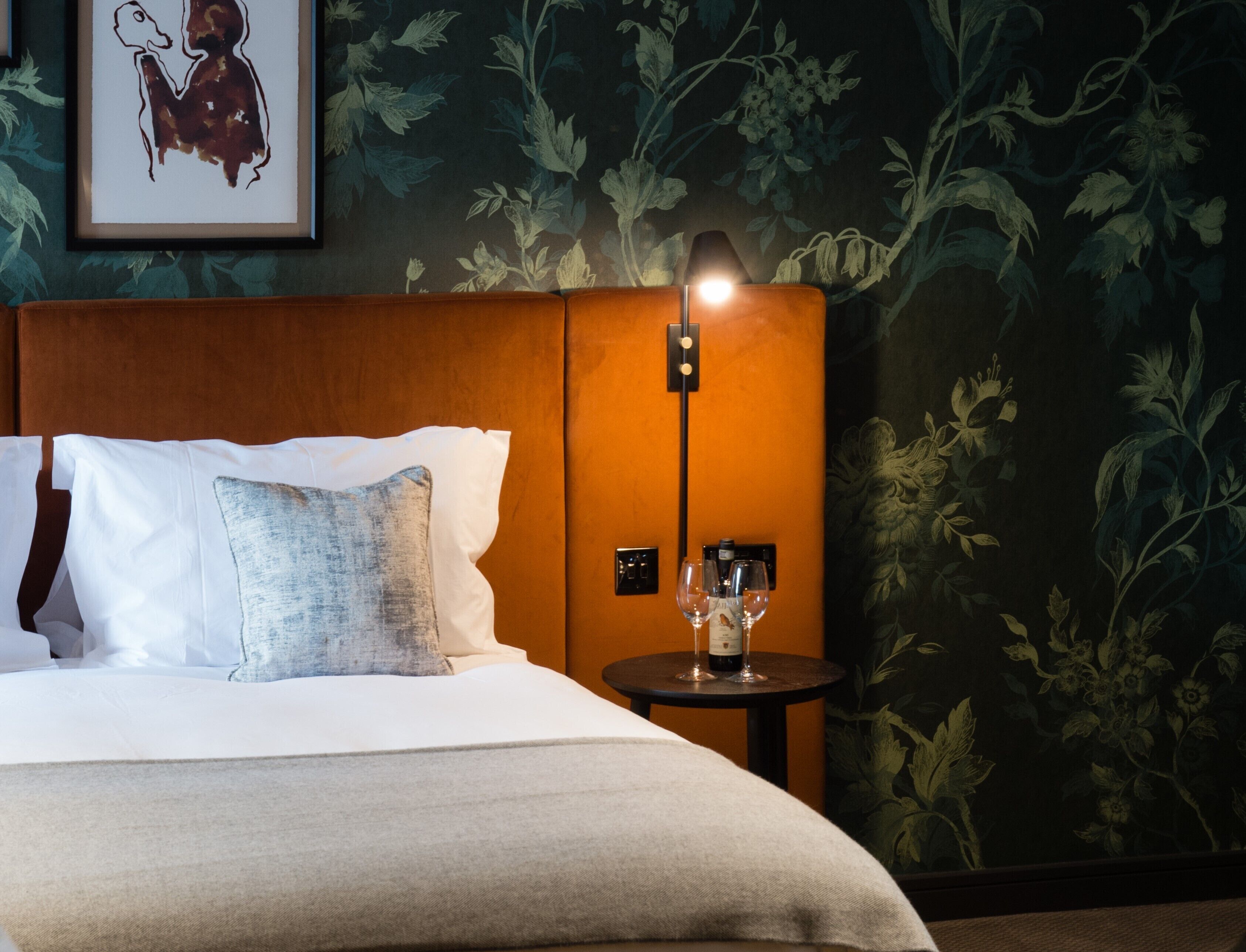 A hotel room with a bed, pillows, a nightstand with a lamp, and a floral wallpaper.