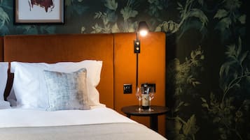 Habitación de hotel con cama, almohadas, mesita de noche y lámpara, y papel tapiz con motivos florales.