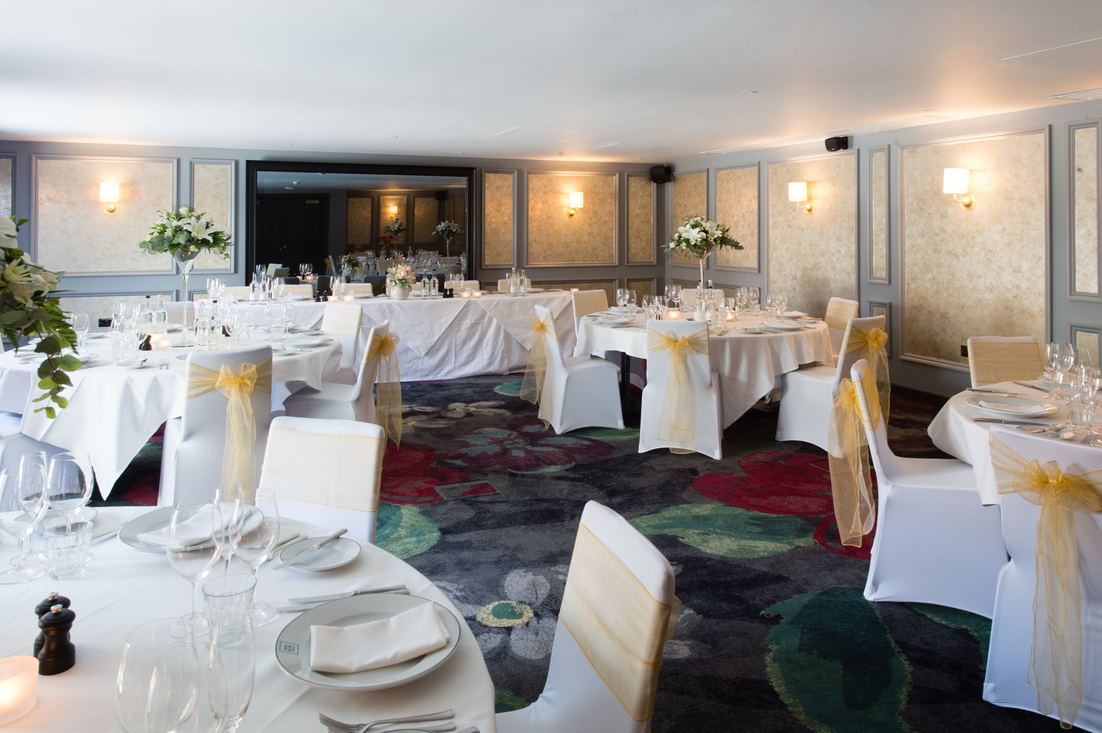 A banquet hall with multiple round tables, each set with white tablecloths, glassware, and chairs with yellow sashes.