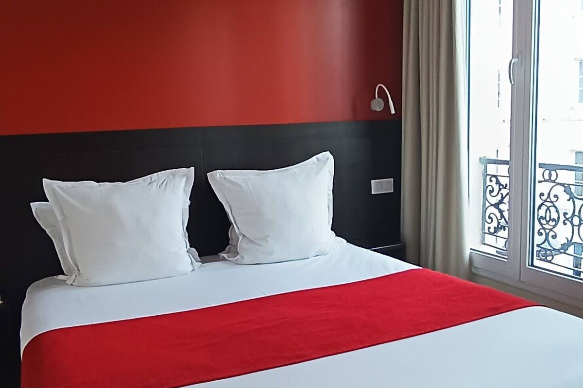 Habitación de hotel con una cama grande, ropa de cama roja y blanca, cabecera negra y vistas a un balcón con barandilla.