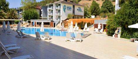 Zona junto a la piscina con sillones de descanso y sombrillas, un edificio con la señalización 'PAM' y 'NOBEL', y una colina al fondo.