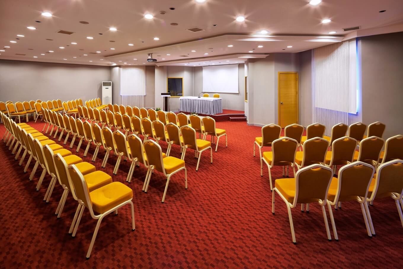 A conference room with rows of yellow chairs, a projector screen, and a table with a white tablecloth.