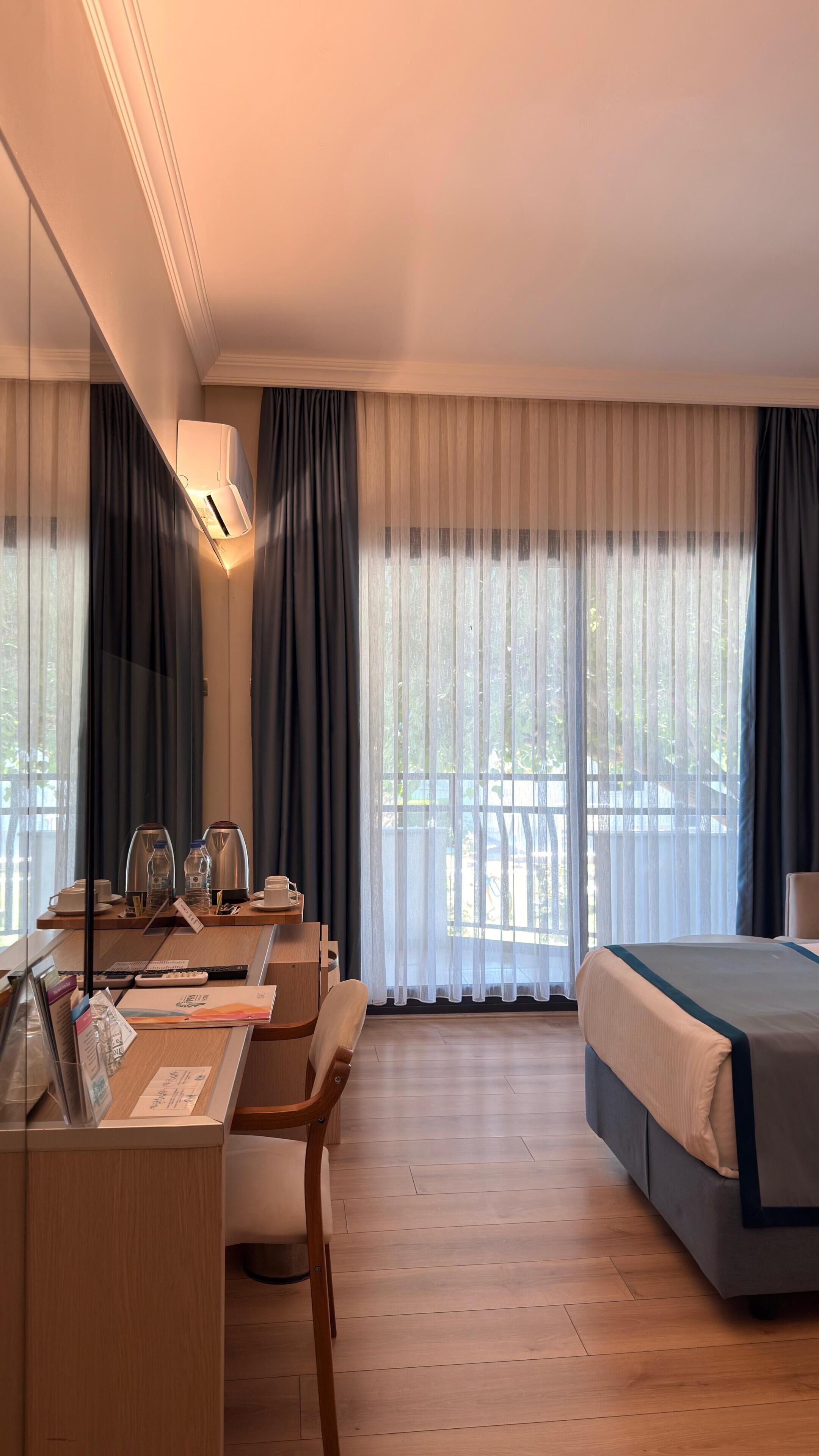 A hotel room with a bed, a desk with chairs, a mirror, an air conditioning unit, and a large window with curtains.