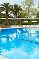 Una piscina con agua azul cristalina, rodeada de sillas de descanso y sombrillas, con palmeras y una zona cubierta al fondo.