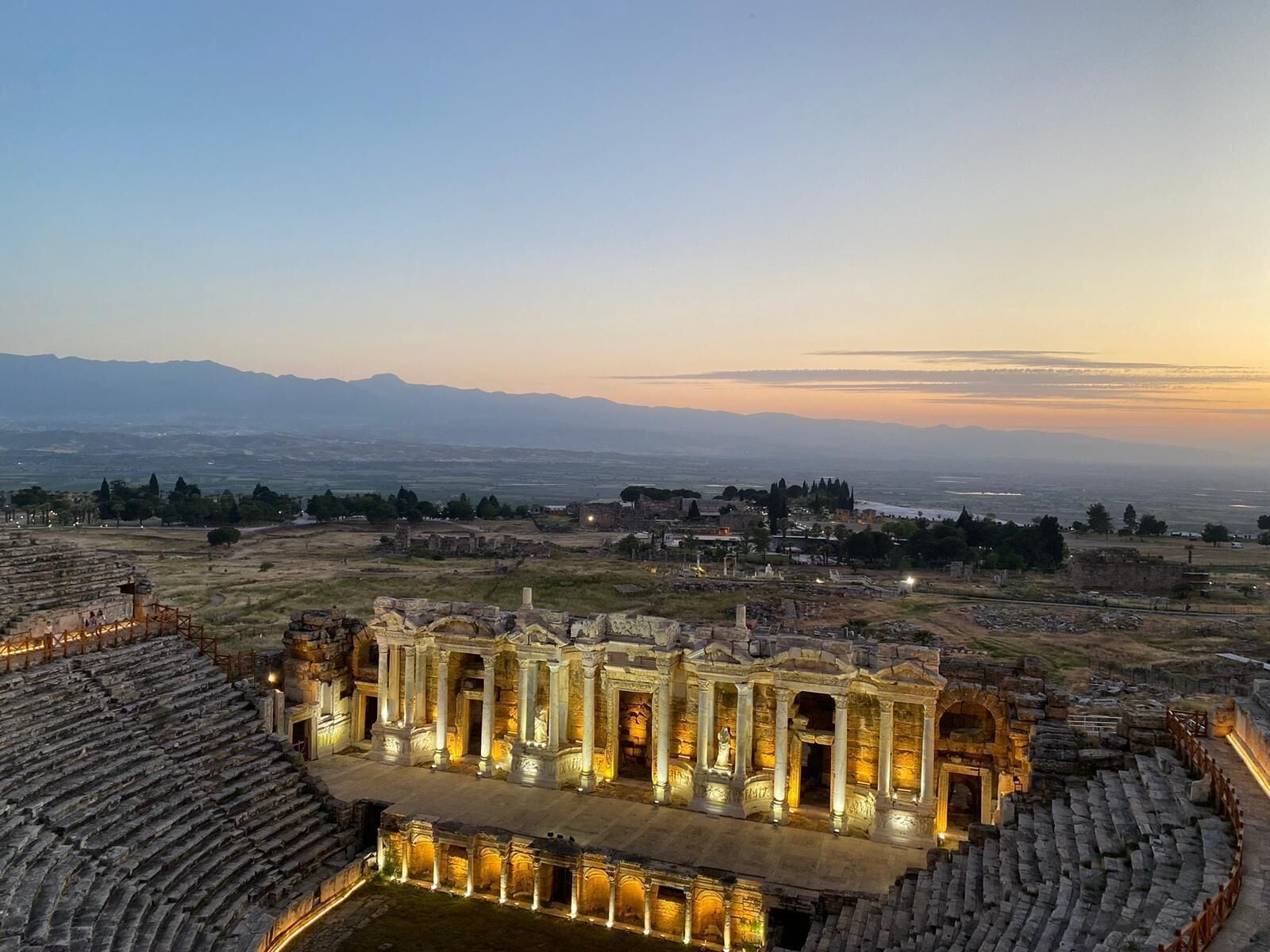 Ein antikes Amphitheater mit Steinsitzen und einer zentralen Bühne, umgeben von Ruinen und einer entfernten Bergsicht.